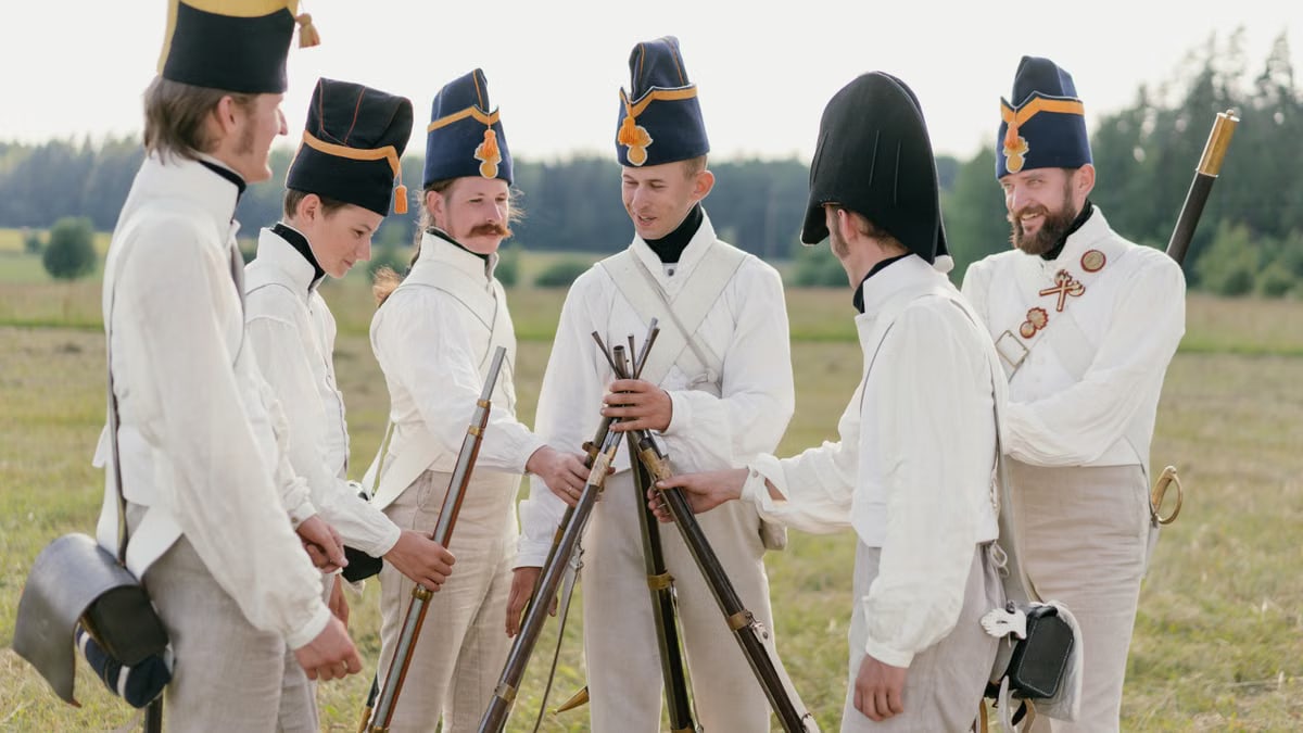 A group of historical reenactors in military uniforms stacking rifles outdoors in a field.